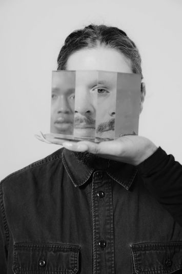 Une personne tient un prisme reflétant plusieurs visages déformés, créant un effet surréaliste. L'arrière-plan est uni et l'image est en noir et blanc.