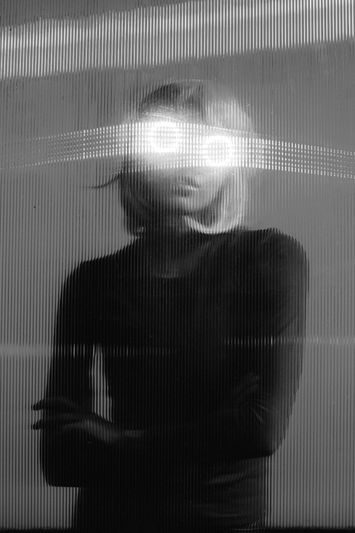 Personne debout derrière un verre texturé avec des yeux brillants et brillants, créant un effet surréaliste et mystérieux en noir et blanc.