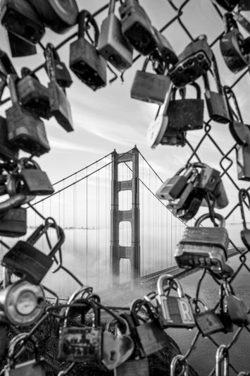 Vue du Golden Gate Bridge encadrée par une clôture en chaîne recouverte de nombreux cadenas, en noir et blanc.