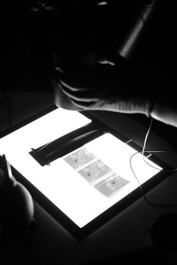Hands holding a camera over a lightbox displaying film negatives, with a dark background.