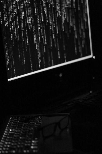 A dimly lit computer screen displays lines of code in a dark room, with a keyboard and glasses visible in the foreground.