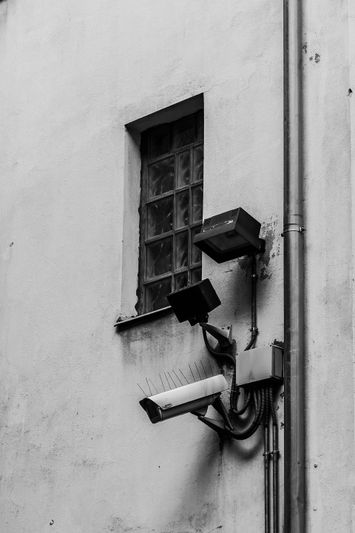 Image en noir et blanc d'un mur avec une petite fenêtre à barreaux, deux caméras de surveillance et un tuyau d'évacuation.