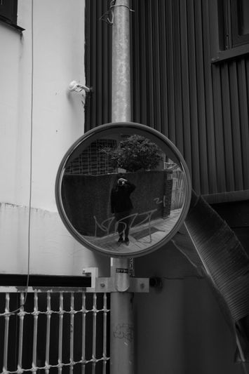 A person in dark clothing is reflected in a round convex mirror attached to a pole, set against an urban backdrop. Black and white photo.
