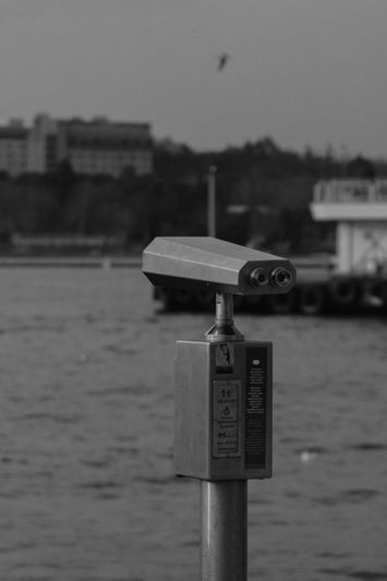Des jumelles à pièces sont installées au bord d'un quai, avec un paysage urbain flou et un oiseau en vol en arrière-plan. Image en noir et blanc.