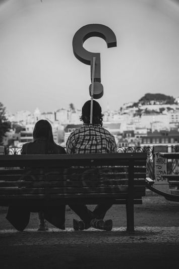 Una pareja está sentada en un banco, de espaldas a la cámara, con un gran signo de interrogación sobre ellos, mirando un paisaje urbano en blanco y negro.