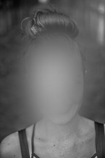 A black-and-white portrait photo of a woman with her hair tied up in a bun, wearing a black tank top, with her face anonymized.