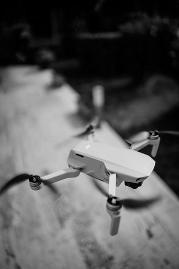 Black and white image of a drone hovering outdoors, with blurred background and sunlight casting shadows.