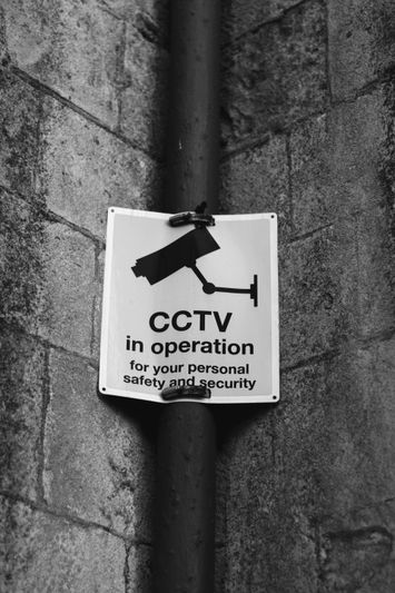 Image en noir et blanc d'un panneau de vidéosurveillance sur un mur de pierre, indiquant « CCTV en fonctionnement pour votre sécurité et protection personnelles. »
