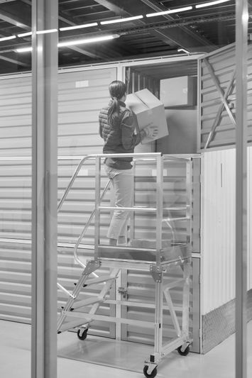 Personne plaçant une boîte dans un box de stockage en hauteur à l’aide d’une échelle mobile.