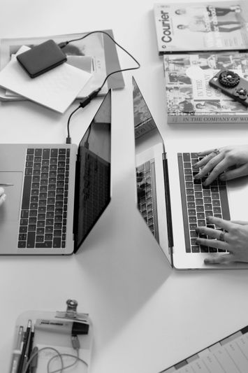 Deux ordinateurs portables sur un bureau, des personnes tapant au clavier, entourées de magazines, d'un carnet et d'appareils électroniques. Image en noir et blanc.