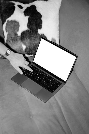 Une personne touche un ordinateur portable avec un écran vide sur un canapé, à côté d'un coussin à motif de vache. Image en noir et blanc.