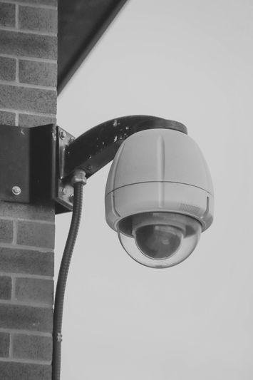 A surveillance camera mounted on a brick wall, featuring a dome-shaped cover and an adjustable arm, captured in black and white.