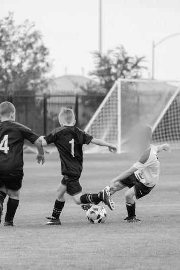 Black and white image of young boys playing soccer, one in white kicking the ball as two opponents in black jerseys attempt to block.