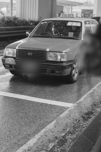 Black and white image of a taxi on a wet road, with a driver inside, under an overpass.