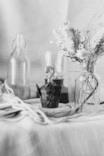 Black and white image of a table with a muffin topped with a question mark candle, surrounded by glass bottles and dried flowers.