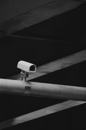 A white security camera mounted on a metal beam, set against a background of intersecting diagonal shadows in grayscale.