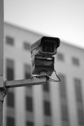 Black and white image of a security camera mounted on a wall, with a blurred building in the background.