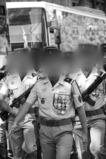 A group of uniformed soldiers with medals, blurred faces, and rifles walk in formation in front of a bus. Black and white photo.