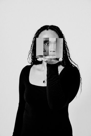 Persona con una camiseta negra, sosteniendo un espejo fragmentado que refleja su rostro sobre un fondo liso. Imagen en blanco y negro.