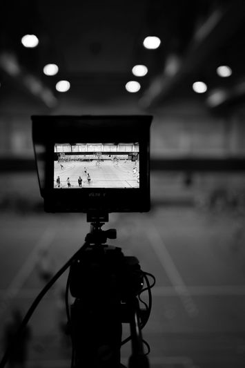 A camera on a tripod captures a sports match in a gymnasium, shown on its screen in black and white.