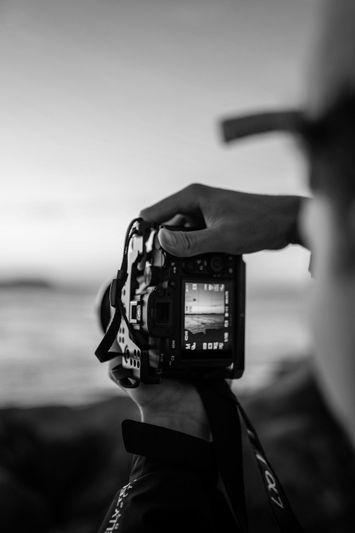 Czarno-białe zdjęcie przedstawiające osobę trzymającą aparat fotograficzny, skupiającą uwagę na morskim krajobrazie wyświetlanym na ekranie aparatu.