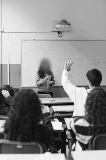 Un maestro está de pie al frente de un aula con una pizarra blanca, mientras un estudiante levanta la mano para hacer una pregunta. Imagen en blanco y negro.