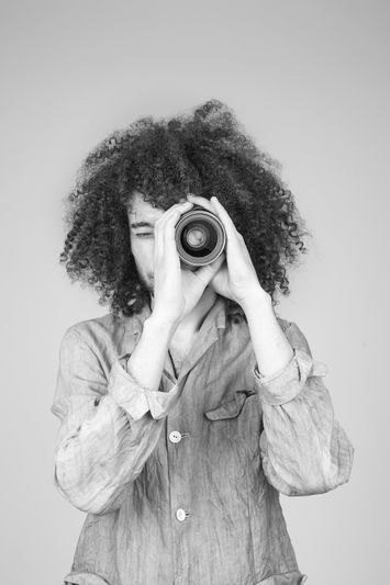 Personne aux cheveux bouclés regardant à travers l'objectif d'un appareil photo tenu comme un télescope, portant une chemise boutonnée sur une photo en niveaux de gris.