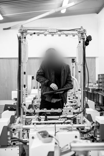 Persona de pie en una estación de trabajo con el rostro borroso, rodeada de maquinaria en un entorno de fábrica. Imagen en blanco y negro.