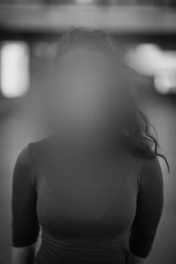 Black-and-white soft-focus portrait of a person from the chest up; face obscured, wavy hair and blurred lights in the background.