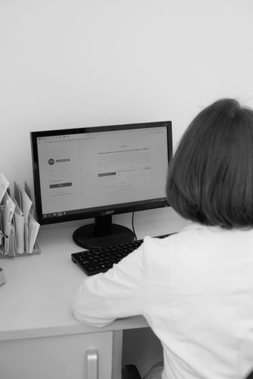 Personne en blouse blanche utilisant un ordinateur à un bureau, avec des documents organisés dans des supports à proximité.