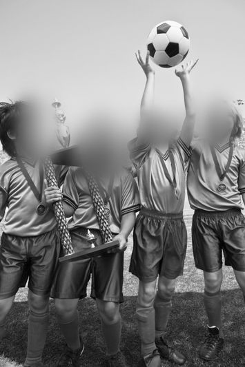 Youth soccer team celebrating with a trophy and medals. One player holds a soccer ball aloft, expressing joy and achievement. Black and white image.