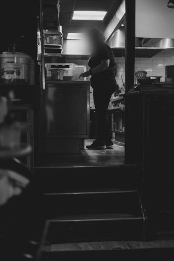 Una persona trabajando en una cocina con poca luz, de pie junto a un mostrador con estantes y equipos visibles a su alrededor.