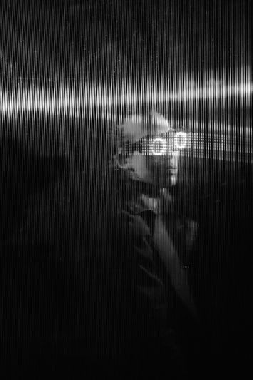 A person wearing glasses is illuminated by a horizontal light beam in a dark, textured setting, creating a futuristic and mysterious atmosphere.