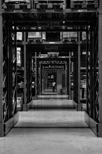 B/W image of a long warehouse aisle with repeating metal shelving and stacked pallets, a single worker visible in the distance.