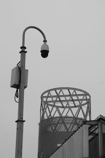 Una imagen en blanco y negro con una cámara de seguridad en un poste y un edificio moderno con una estructura geométrica de celosía contra un cielo gris.