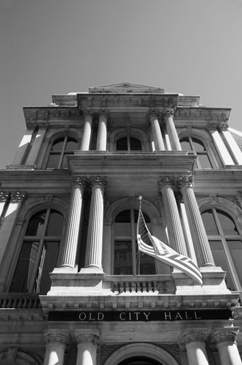 Edificio histórico con altas columnas y ventanas con arcos. Una bandera estadounidense cuelga de manera destacada. Un cartel dice 'Antiguo Ayuntamiento'. Foto en blanco y negro.