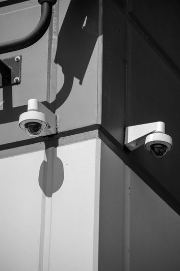 Deux caméras de sécurité montées sur un coin d'immeuble, projetant des ombres sur le mur en noir et blanc.