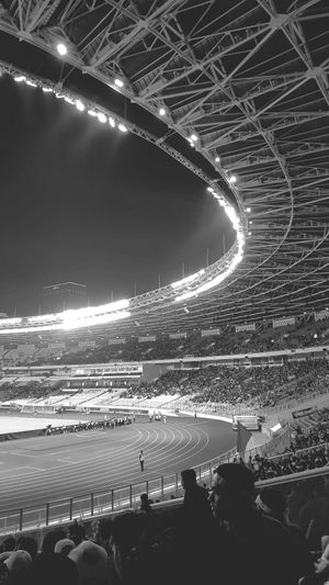 Black and white photo of a large stadium at night, with bright lights illuminating the track and field, and a crowd in the stands.