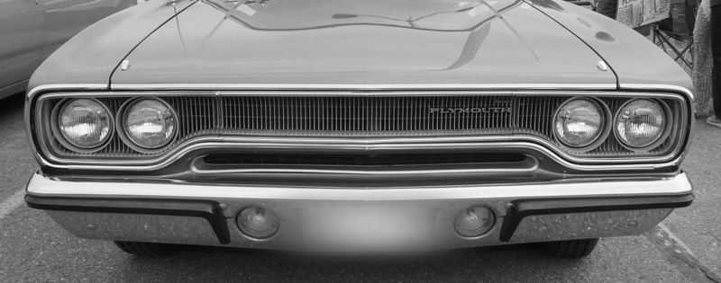 black and white photo of the front of a 'Plymouth' car has a blurred license plate