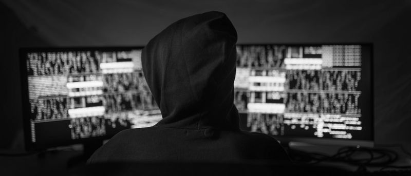 Una foto en blanco y negro que muestra a una persona con capucha sentada de espaldas a nosotros, mirando dos pantallas de computadora.