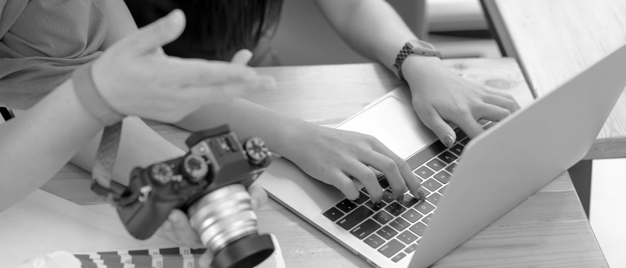Persona escribiendo en una computadora portátil mientras otra sostiene una cámara, discutiendo algo en una mesa de madera.