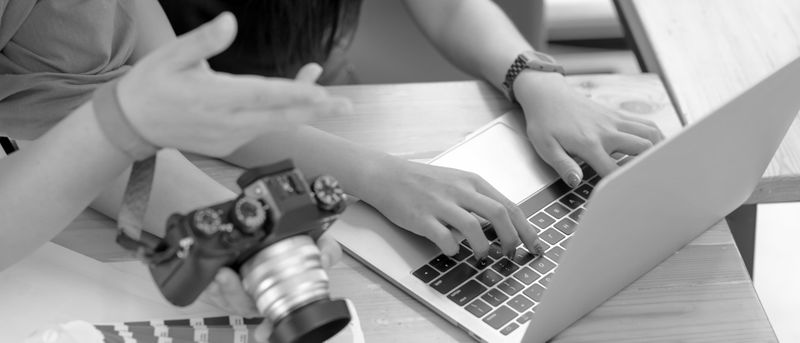 Person typing on a laptop while another holds a camera, discussing something at a wooden table.