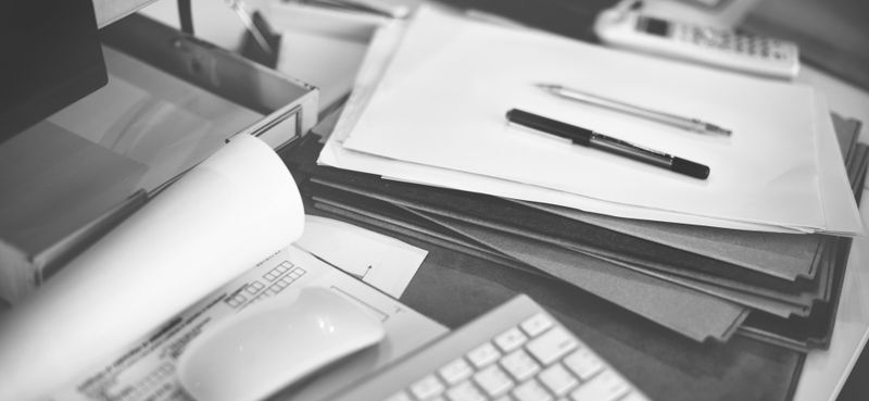 A black-and-white photo of a cluttered desk with papers, a pen, a keyboard, a mouse, and a phone.