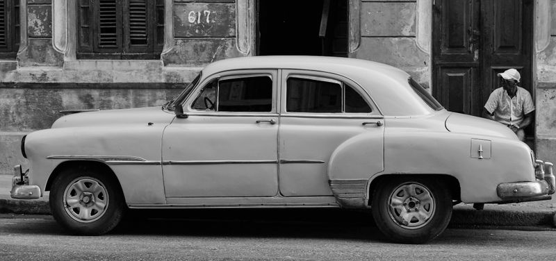 Coche antiguo estacionado en una calle frente a un edificio viejo; una persona con un sombrero se apoya en la pared en el fondo. Blanco y negro.