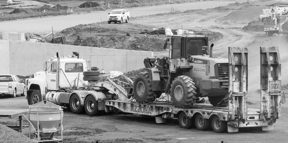 Ciężarówka z platformą transportuje duży pojazd budowlany na placu budowy, w tle widać stosy ziemi i pojazdy. Czarno-biały obraz.