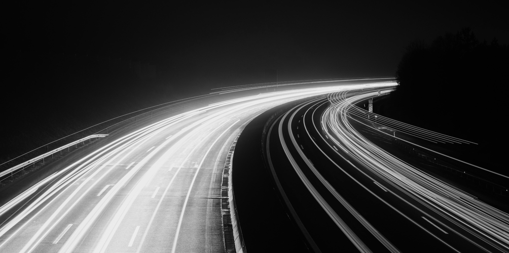Schwarzweißfoto einer Autobahn bei Nacht, das durch lange Belichtungszeit Lichtspuren von Fahrzeugen zeigt, die helle, geschwungene Linien vor dem dunklen Himmel erzeugen.