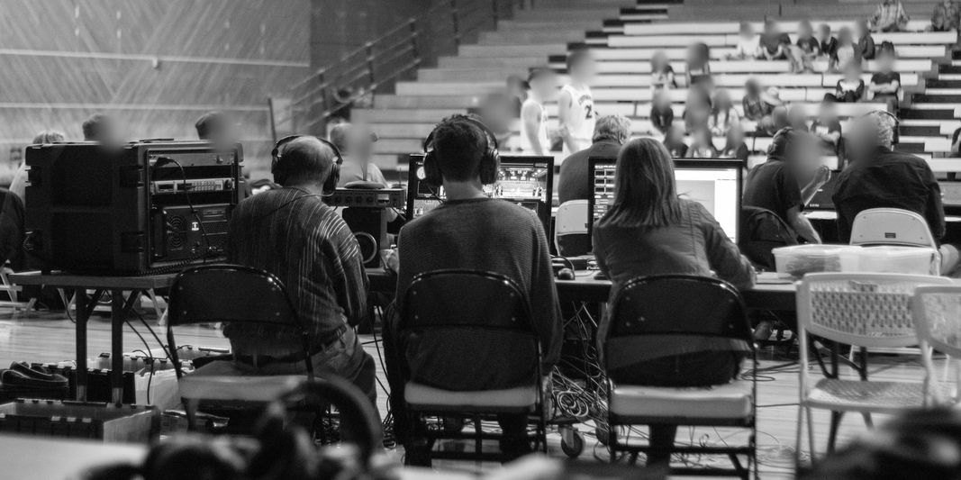 Three people seated at a control table with audio and video equipment in a gymnasium, facing a blurred audience.