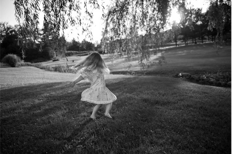 A young girl in a dress twirls on a grassy field under a tree, with sunlight filtering through the branches in a serene landscape.