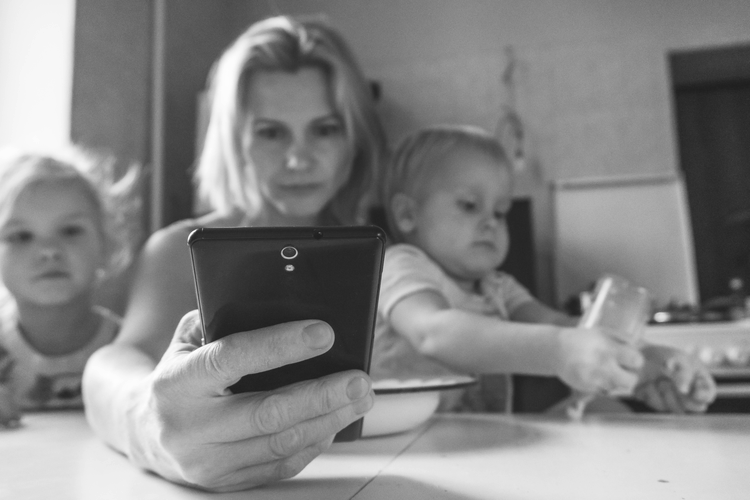 Kobieta patrzy na swój telefon, podczas gdy dwoje dzieci siedzi obok niej przy stole, jedno trzyma butelkę. Sceneria wydaje się być kuchnią.