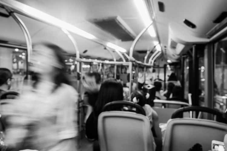 Image floue en noir et blanc de personnes assises et debout à l'intérieur d'un bus, avec des sièges visibles et des poignées de porte au-dessus de la tête.